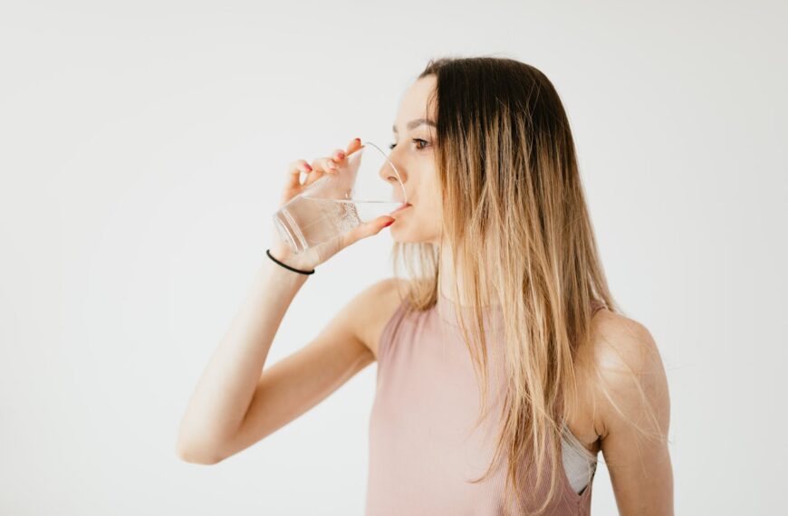 Young thirsty fit female with long hair in sportswear drinking water while recreating after workout